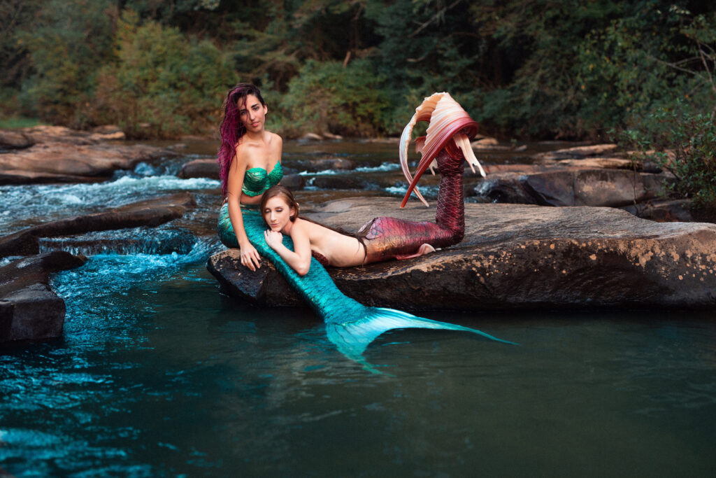 Mermaid Jules sits on the edge near a stream, a mermaid friend sleeps in her lap.