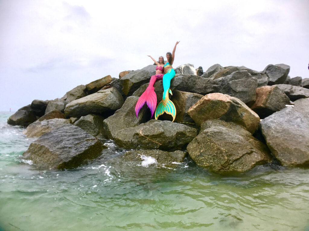 Two smiling mermaids throw their arms in the air, sitting a top boulders in the ocean.