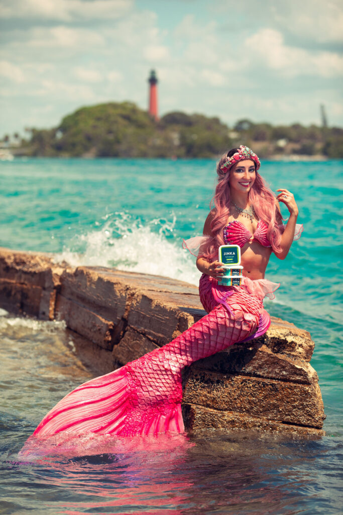 Mermaid Jules sits with her Jinka Tuna Spread alternative snack as waves splash around her.
