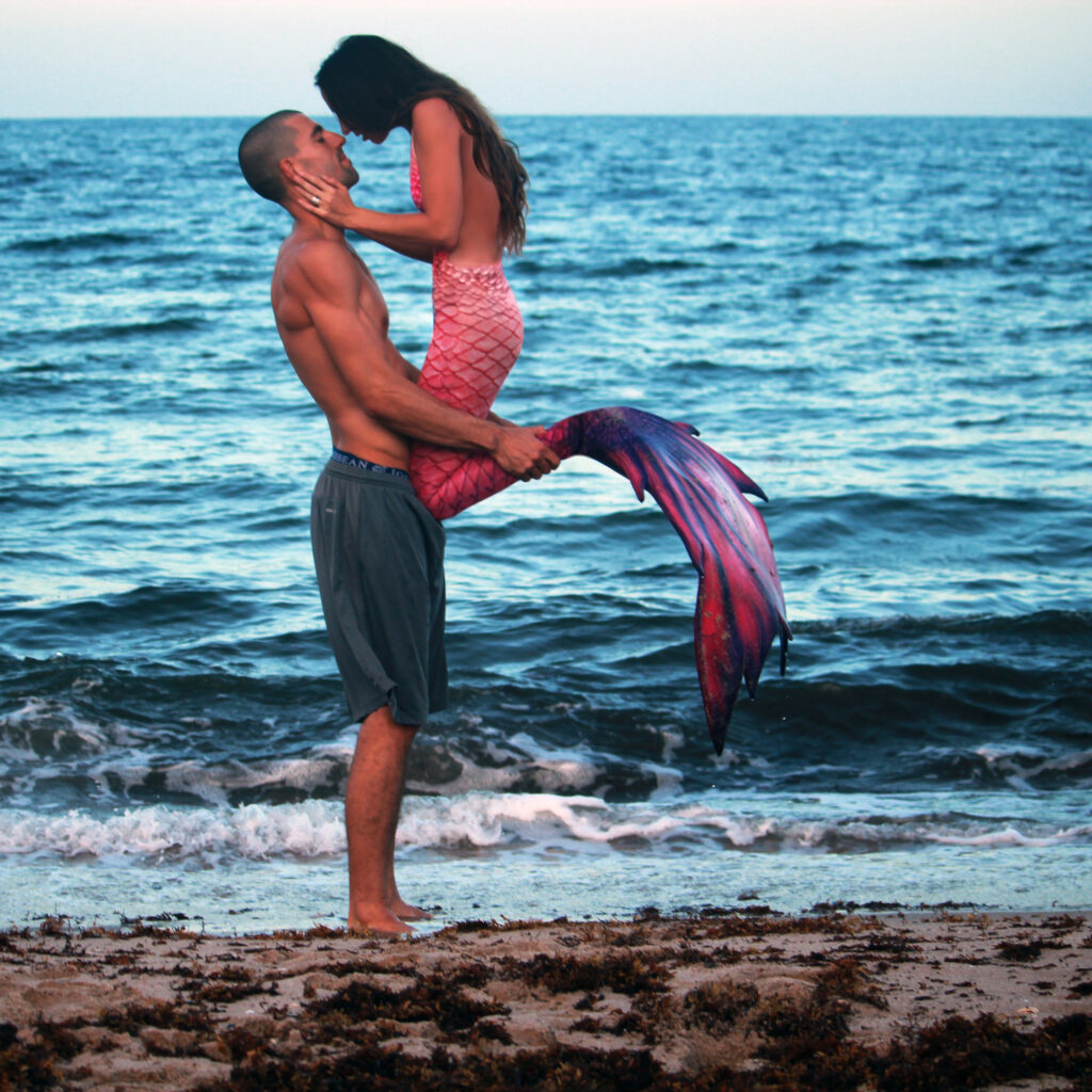 A pink mermaid leans down to kiss the man who is lifting her up, their faces silhouetted by the ocean.