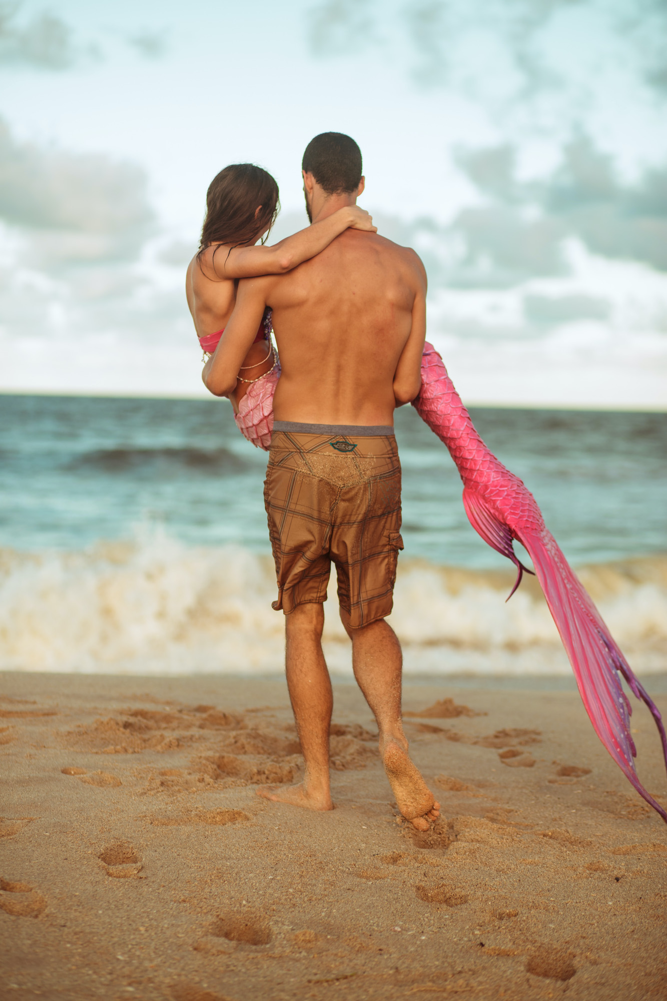 A man on the beach walks towards the crashing waves carrying a pink mermaid in his arms.