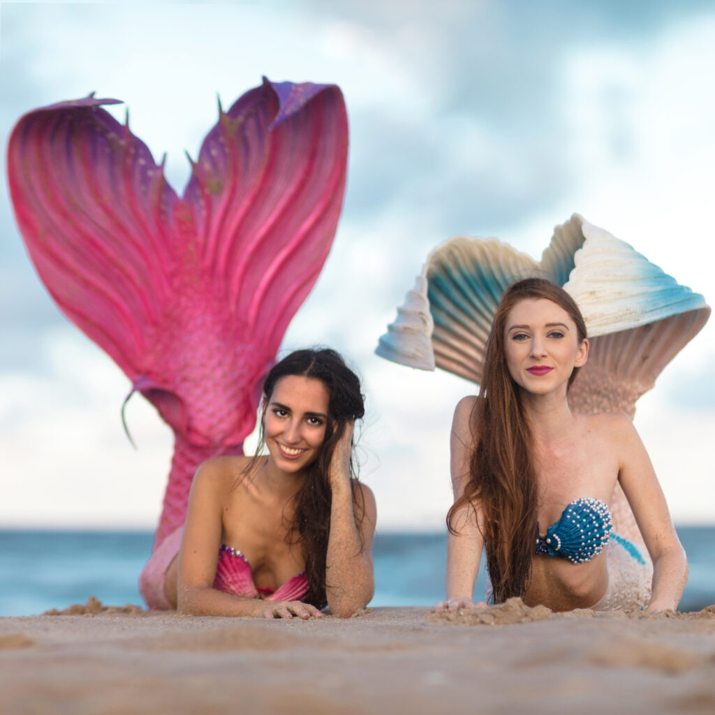 Two mermaids on a beach lay on their bellies looking at you with their flukes in the air.