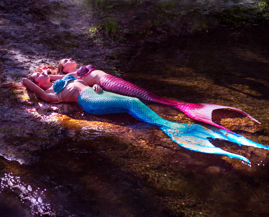 Two mermaids lay out on the edge of a stream.