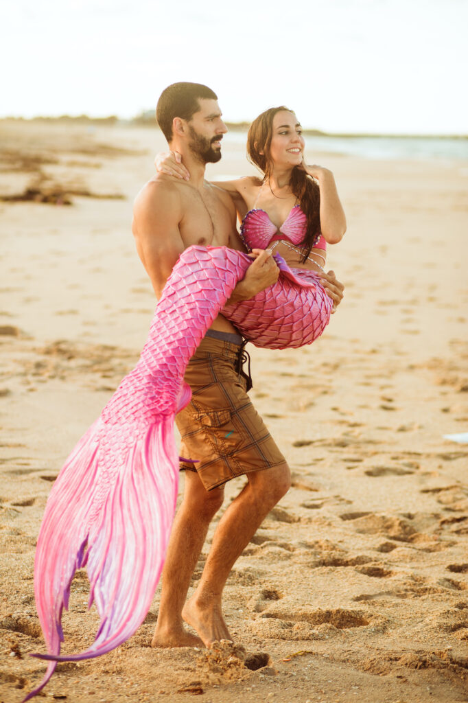 A man on the beach walks towards the sea carrying a pink mermaid in his arms.