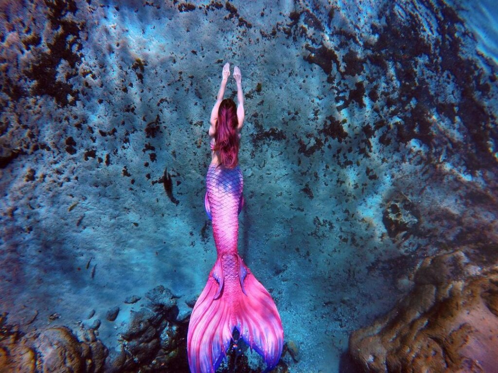 Mermaid Jules swims in the deep blue; her pink tail pops bright against the blue sea.