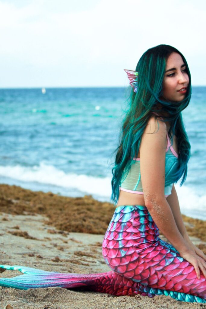 Blue hair wraps around a mermaid's face on the windy shore of the beach,as she looks out over the horizon.