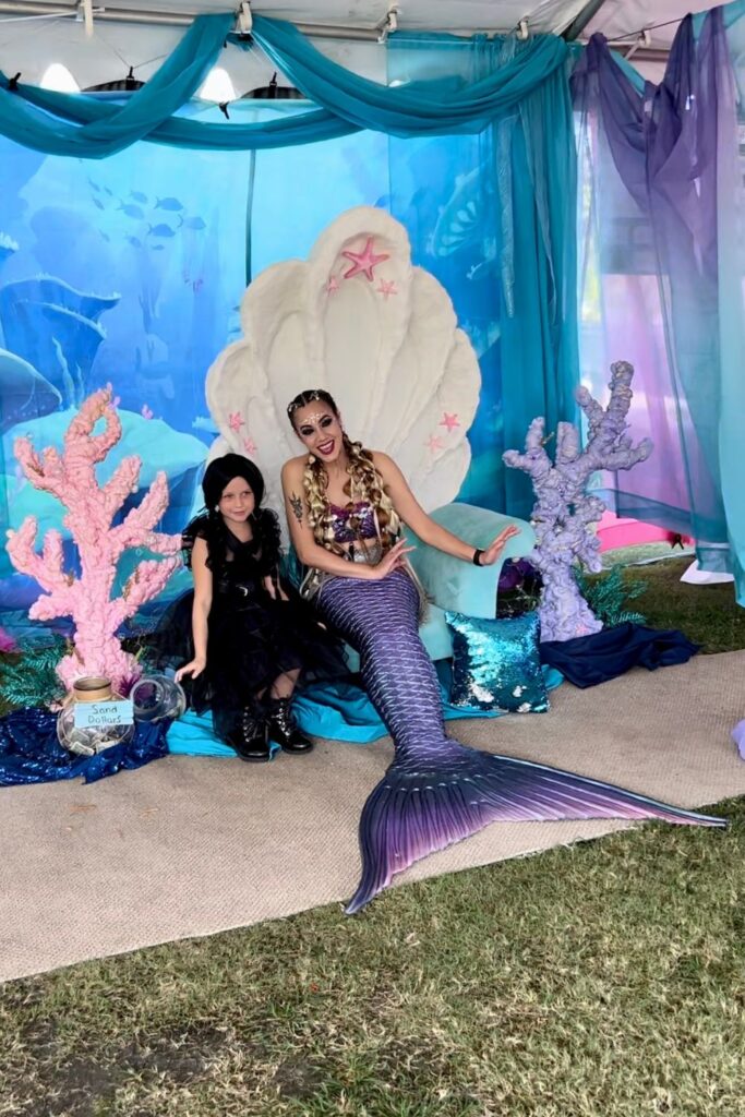 Wednesday Adams costume poses next to mermaid in clam throne